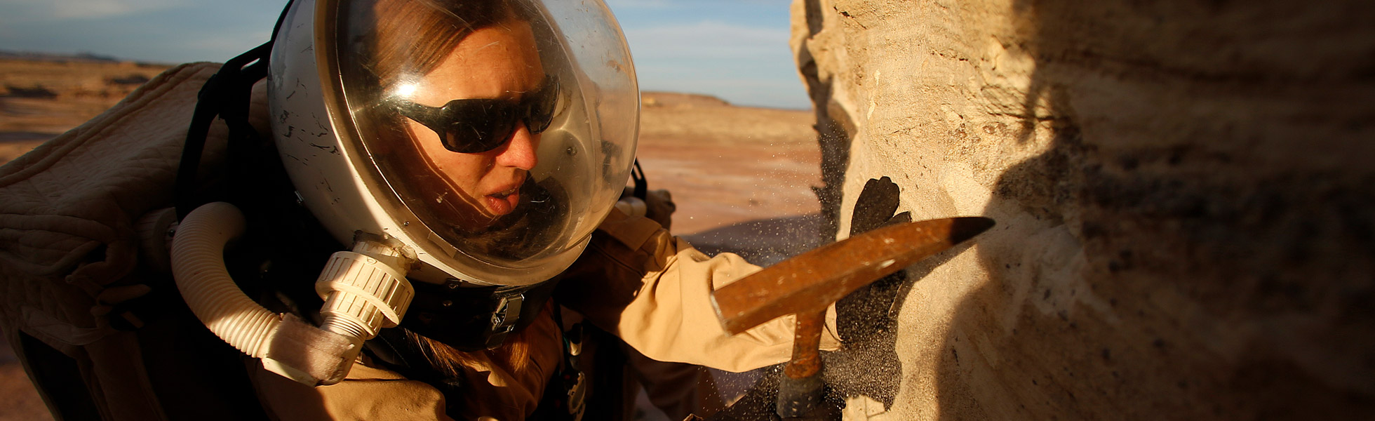 A photo of a woman in a space suit digging into a rock with a small hammer.